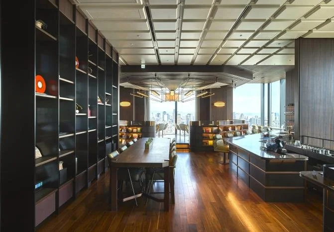 Modern lounge area with large windows, wooden floors, dark shelves, and seating, overlooking a city skyline.