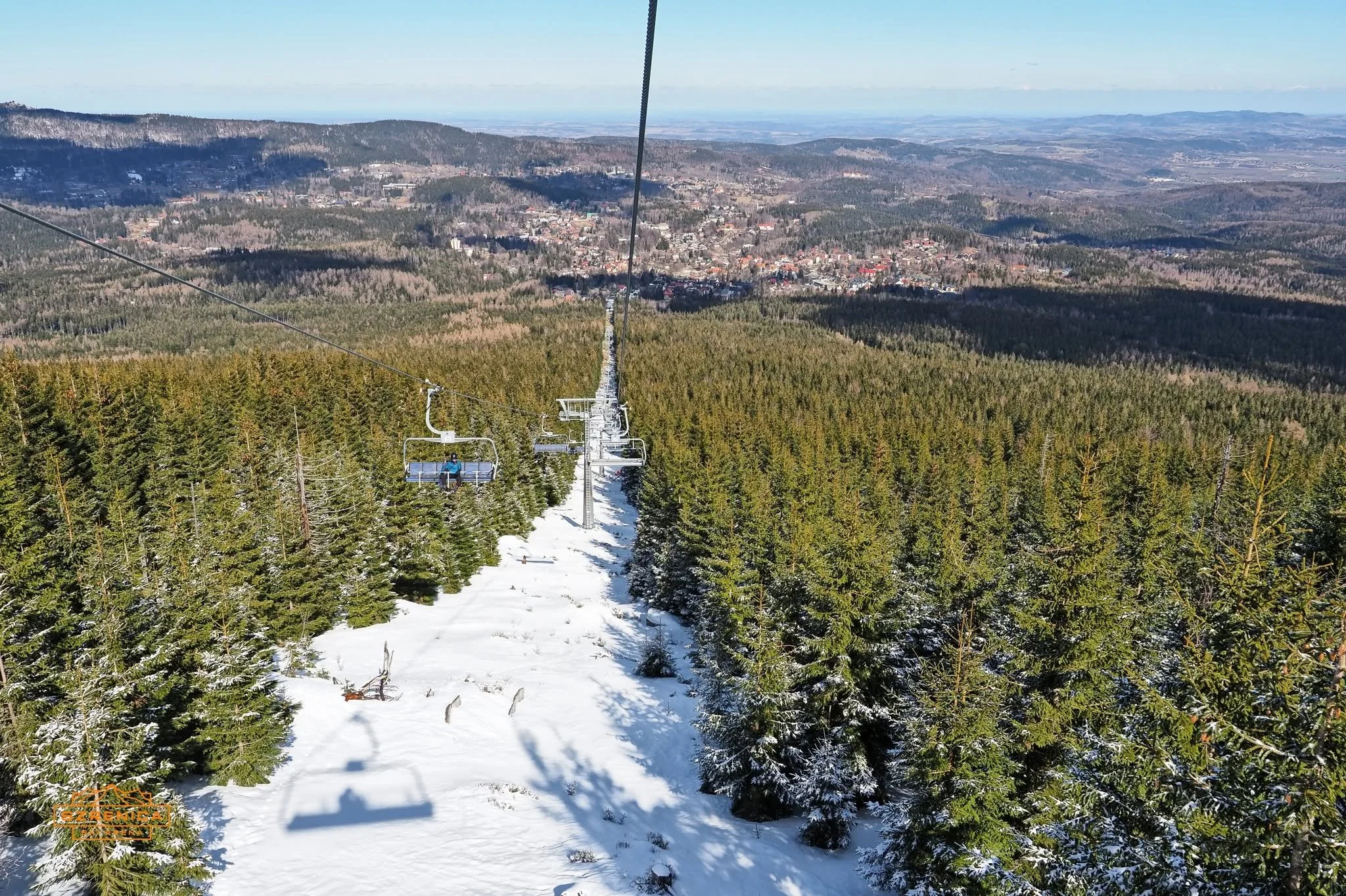 View from a ski lift over a snowy forest with a small town in the distance and mountains in the background.