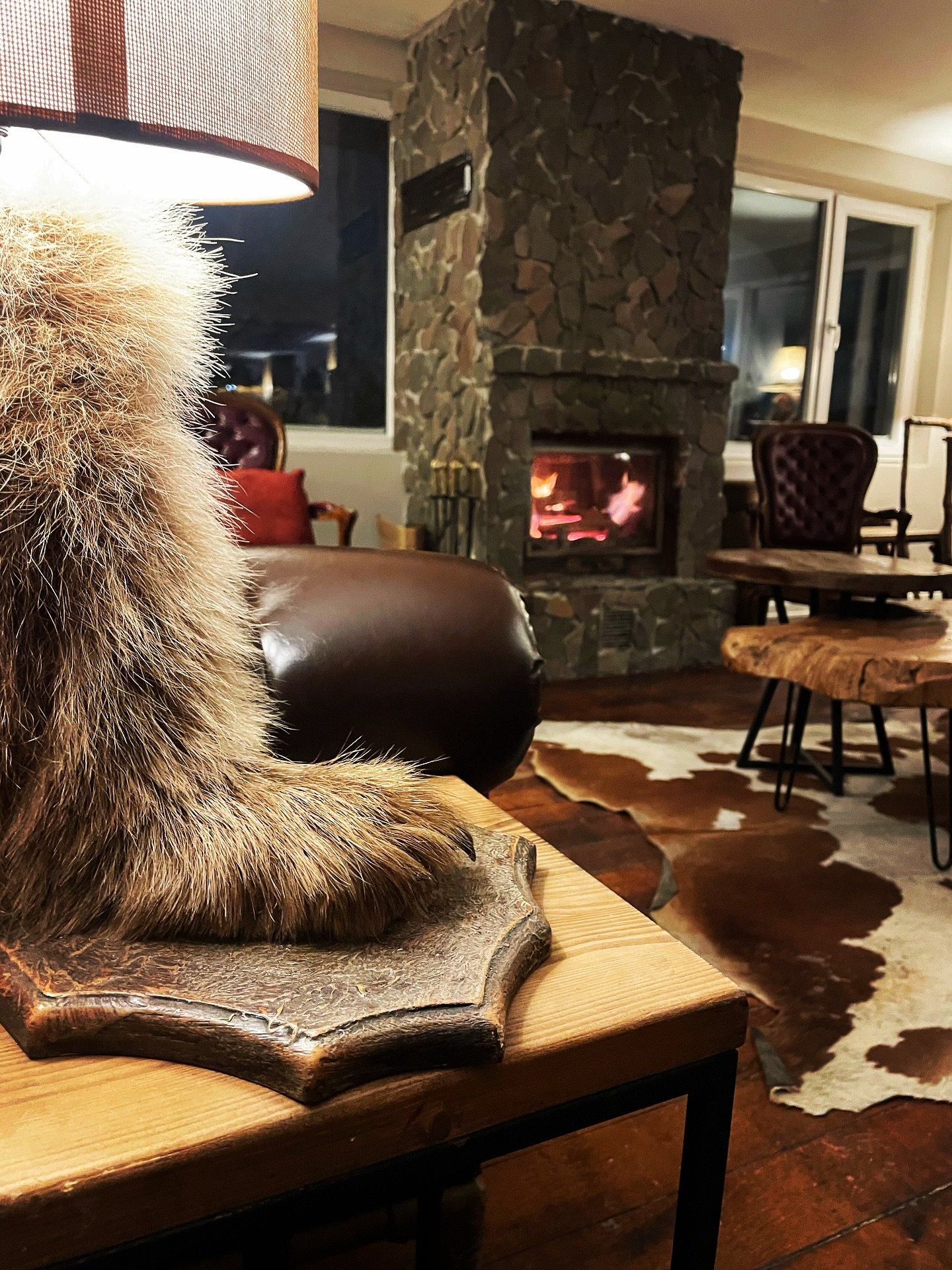 A cozy living room with a stone fireplace lit with a fire, surrounded by vintage chairs and wooden tables, with a cowhide rug on dark wooden flooring.