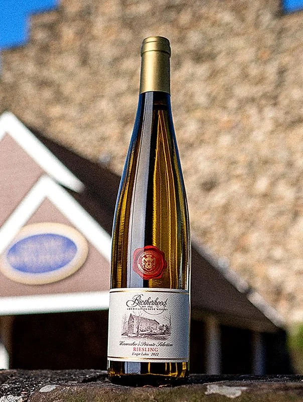 A tall bottle of Brotherhood Riesling wine with a gold cap, placed outdoors on a stone surface, with a house and a brick wall in the background.