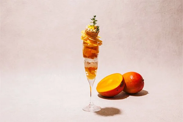 A dessert in a tall glass topped with mango slices, whipped cream, and garnished with a sprig of herbs, next to a whole mango and a mango half.