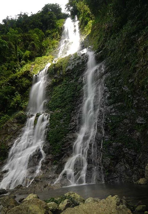Tall waterfall cascading down a rocky hillside surrounded by green trees and foliage.