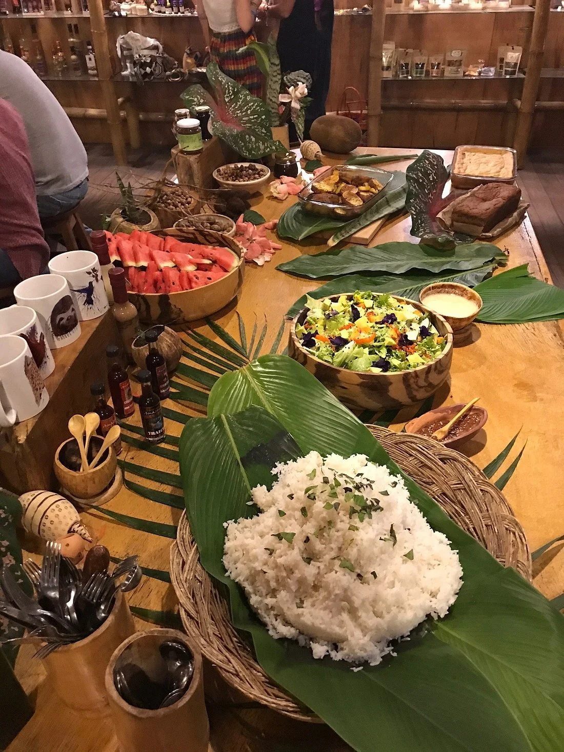 A table set with various foods, including a large bowl of cooked rice with green herbs on top, a bowl of salad, slices of watermelon, and several other dishes, surrounded by large green leaves and small condiment bottles.