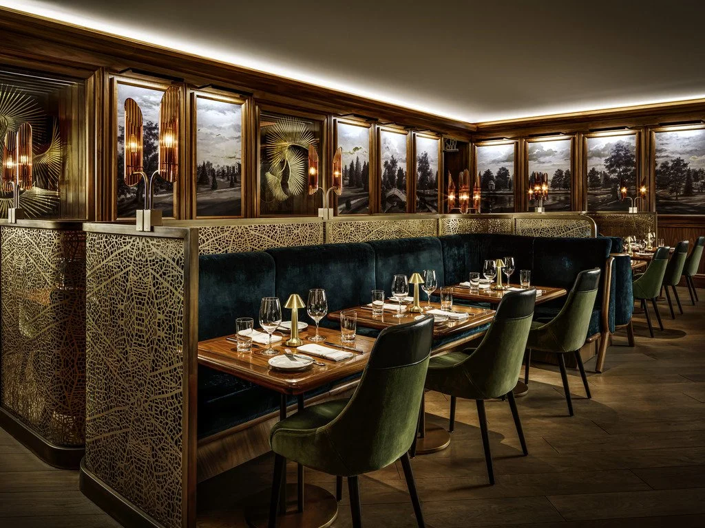 Elegant restaurant with dark green velvet seating, wooden tables set with wine glasses, water glasses, napkins, and candlestick lamps. The space features large panoramic windows with scenic outdoor views, framed by wooden paneling, and warm ambient l