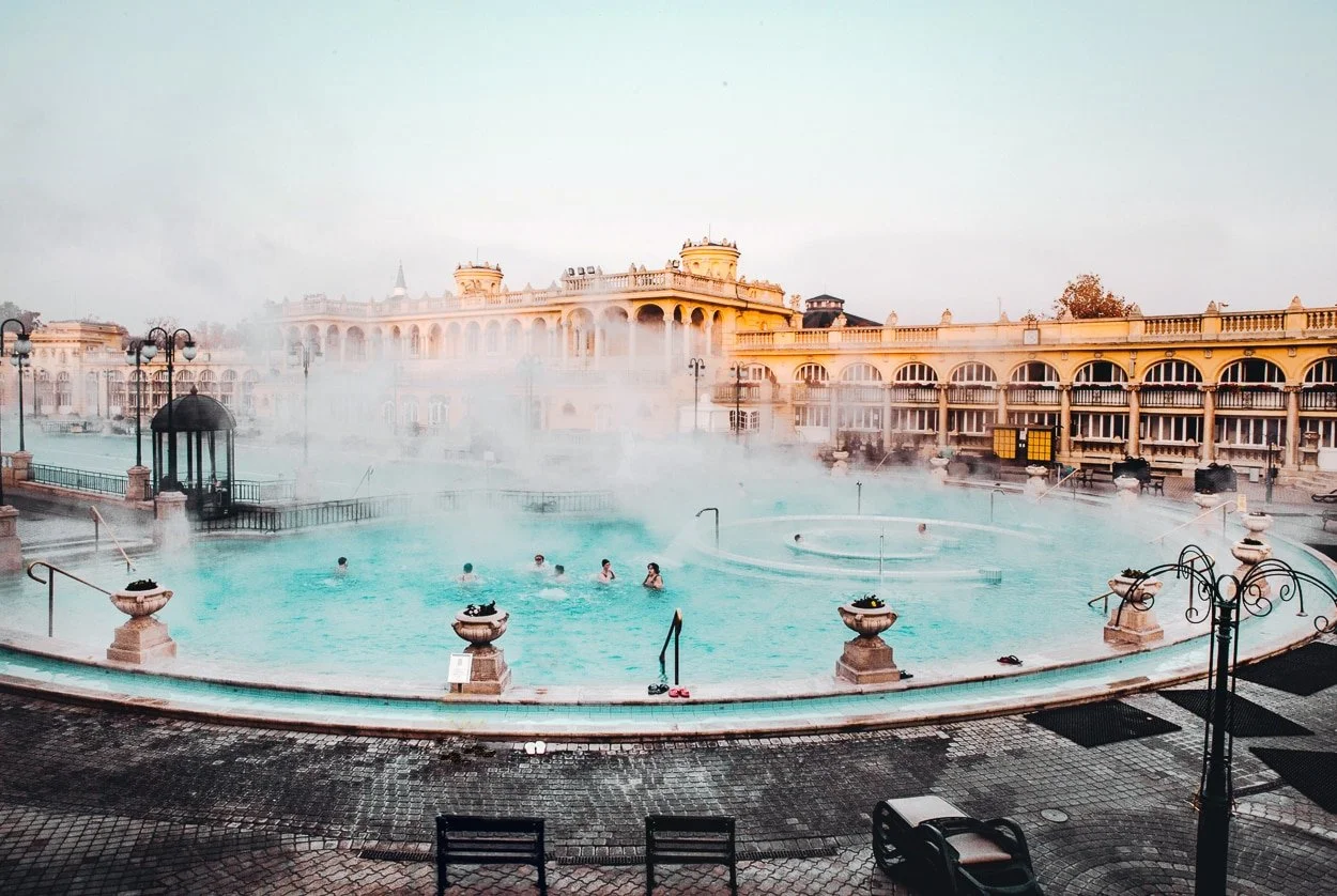 Outdoor hot spring pool with steam rising, surrounded by ornate architecture and street lamps, with people relaxing in the water.