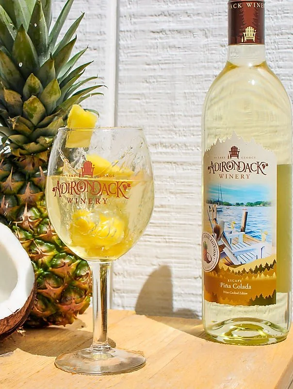 A glass of Pina Colada wine with pineapple chunks and a straw, next to a bottle of Adirondack Winery Pina Colada wine, with a fresh pineapple and a coconut in the background.
