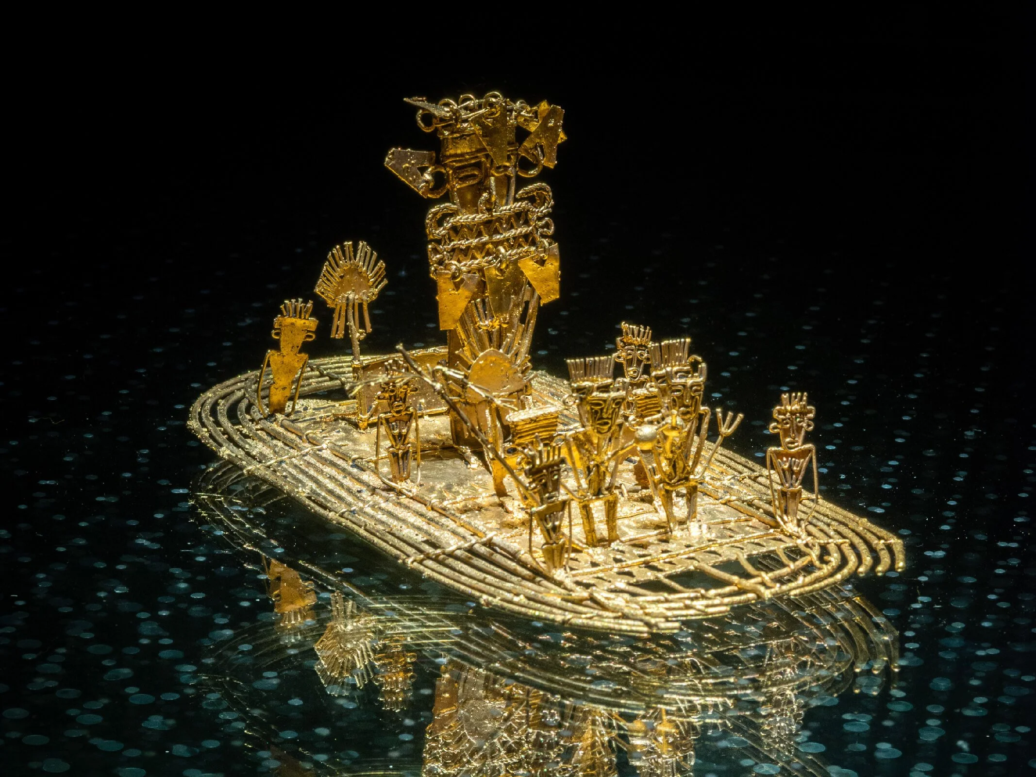 A detailed close-up of a gold jewelry miniature sculpture depicting figures and structures, placed on a reflective surface with a black background.