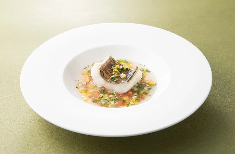 A plated serving of fish, possibly cod or haddock, with a crispy skin, resting on a bed of herbs and diced vegetables in a light, clear broth, presented on a white round plate with a green background.