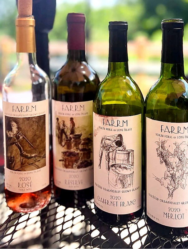 Four bottles of wine with farm-themed labels on a metal surface outdoors. The bottles are labeled Rose, Reserve, Cabernet Franc, and Merlot from 2020.
