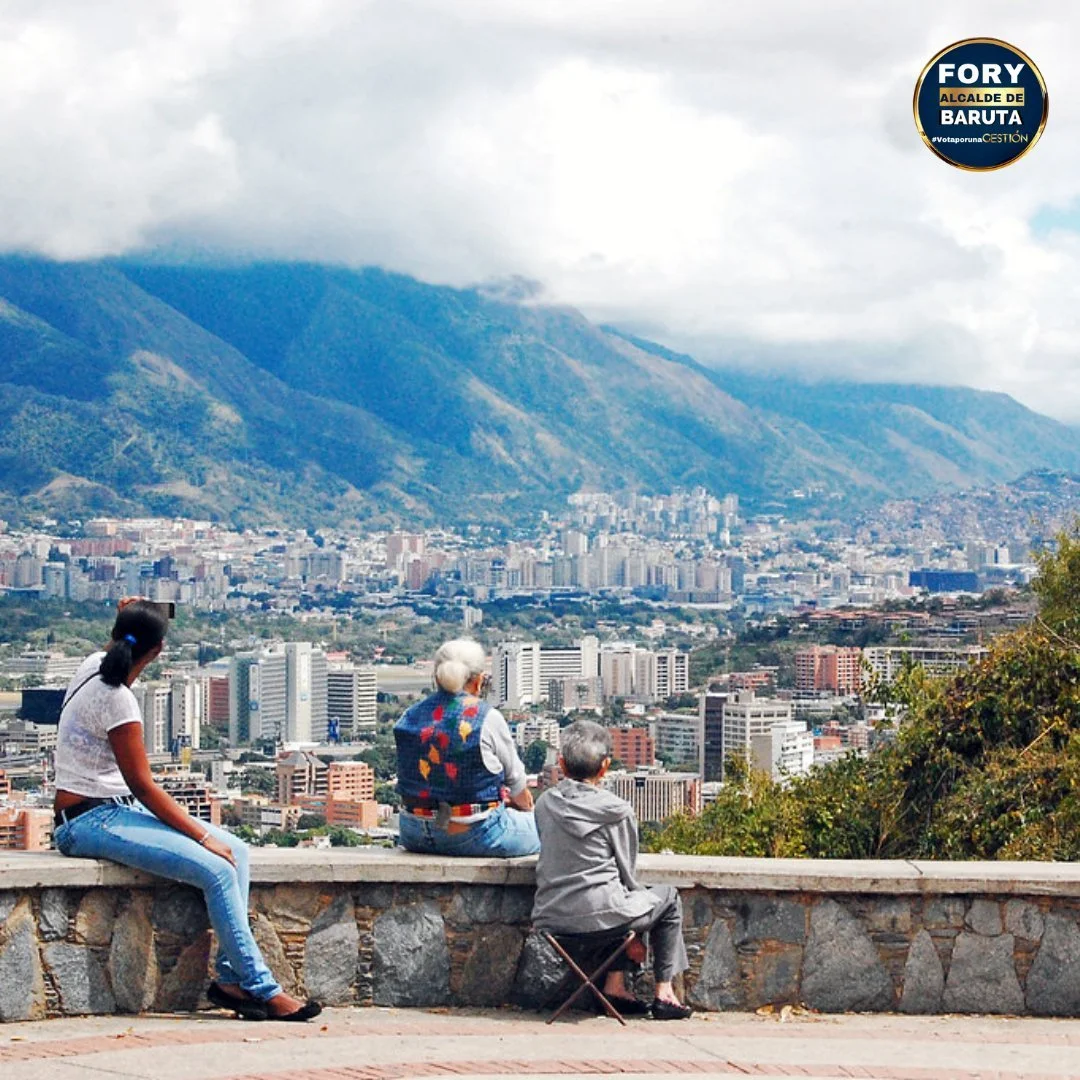 Three people sitting on a stone wall overlooking a city with tall buildings and mountains in the background. One person is a young woman, and two are elderly women. The elderly woman on the right has short gray hair and is sitting on a small stool, w