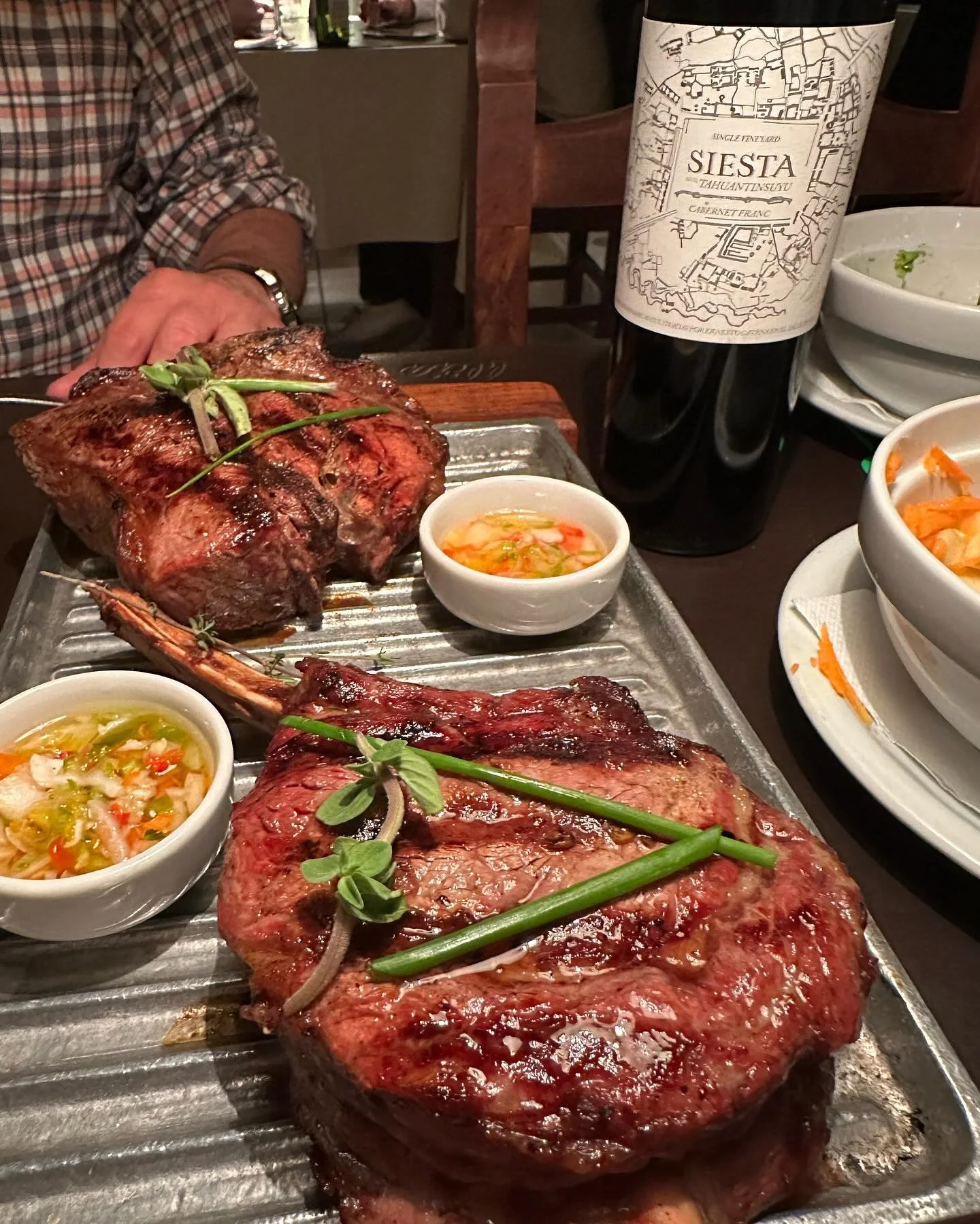 A table featuring two large cooked steaks garnished with green herbs and onions, surrounded by small bowls of vegetables and a bottle of Siesta wine.