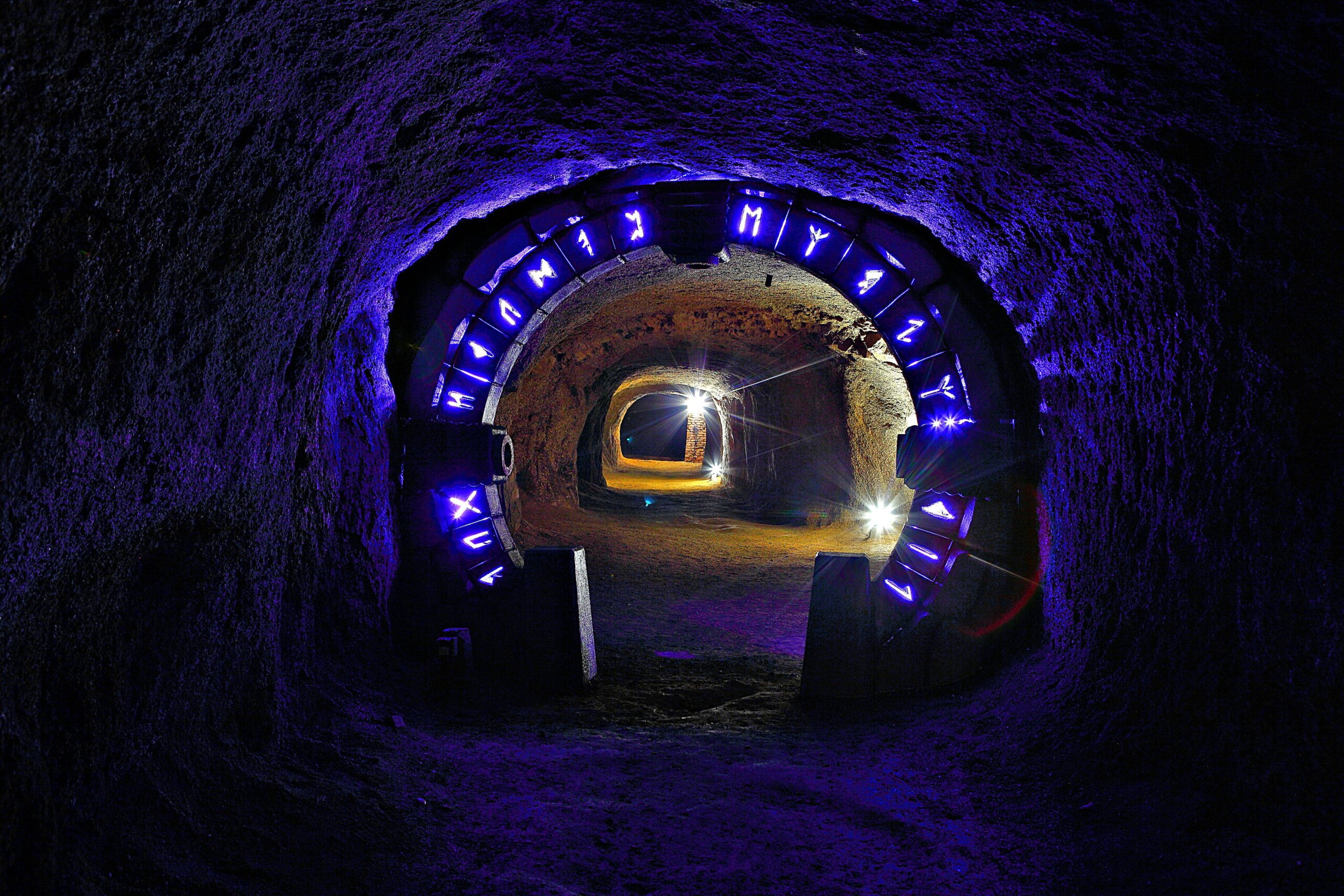 Underground tunnel with pink and purple lighting, a circular metal sign with illuminated letters, and a bright light at the far end of the tunnel.