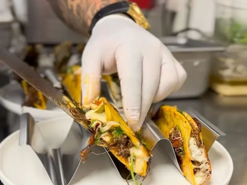 Person wearing gloves preparing baked taquitos with cheese and meat filling in a kitchen.