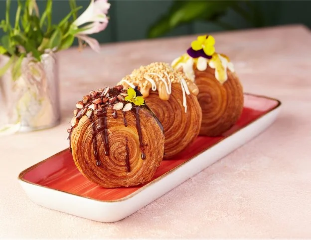 Three flaky, swirled pastries with icing and toppings on a rectangular red plate, with a flower in a vase in the background.