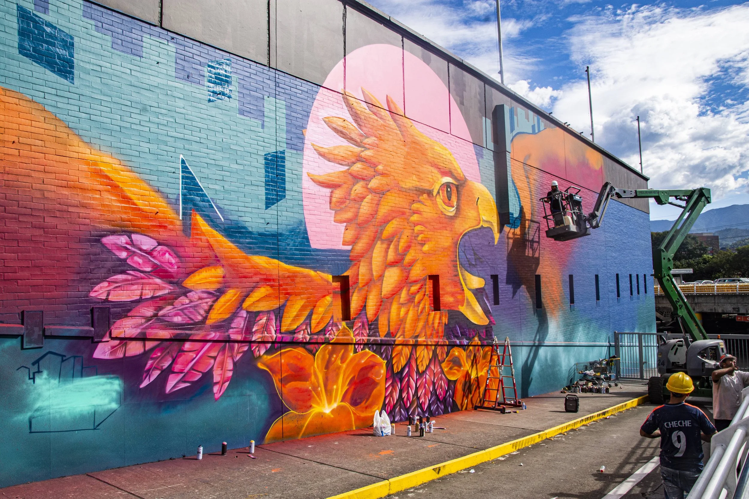 A large colorful mural of a lion's head being painted on a building wall. A worker in a bucket lift is working on the mural, while others are on the ground observing.
