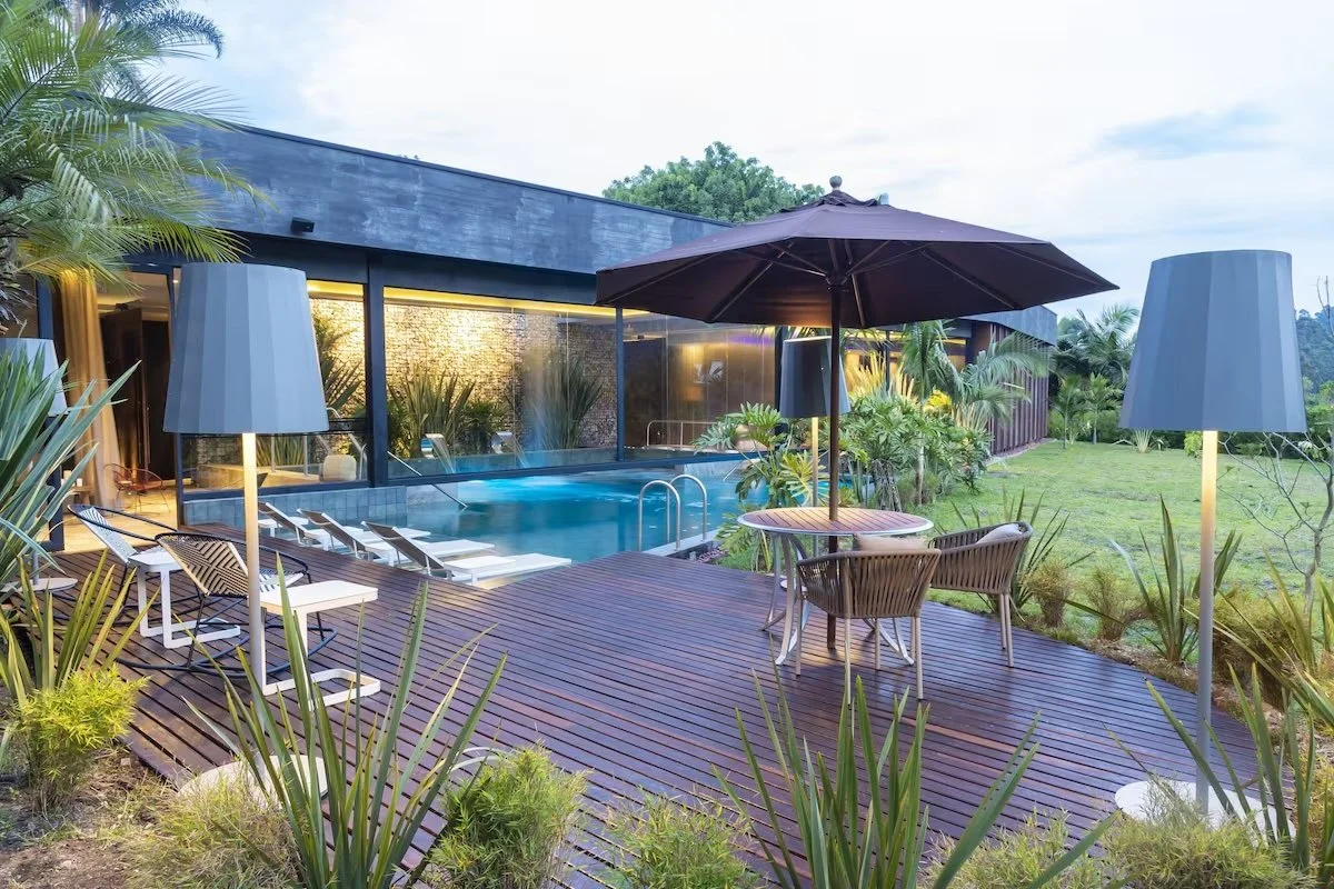 Outdoor pool area with lounge chairs, tables, umbrellas, and surrounding greenery at a modern house or resort.