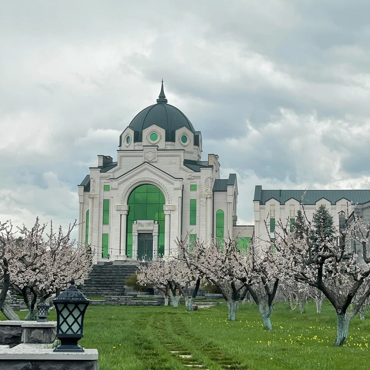 A large white building with green-tinted windows and a dark dome in the center, surrounded by blossoming trees and a cloudy sky.