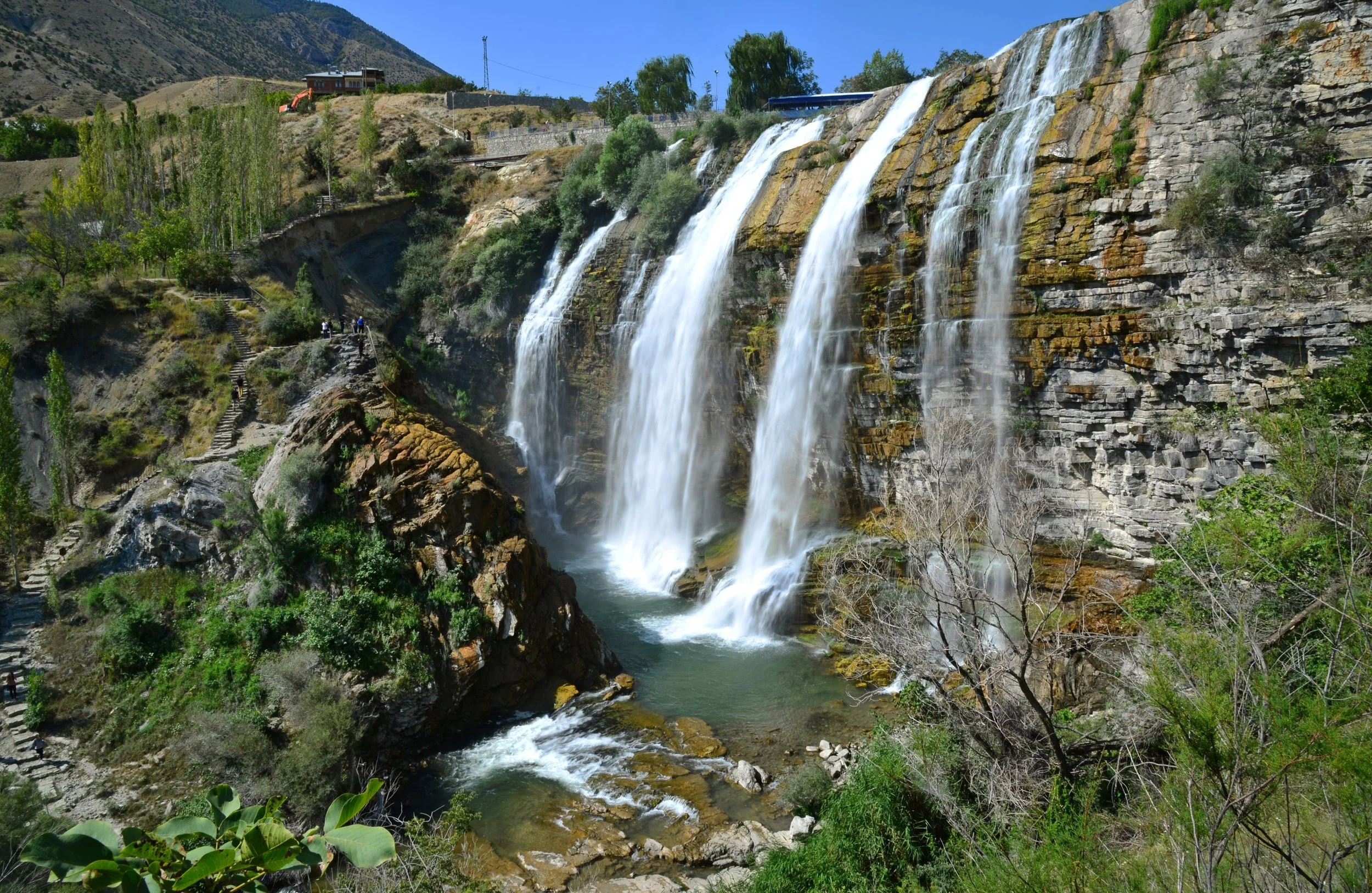 Waterfall flowing over rocky cliffs with lush green trees and steps on the left side leading down to the water, under a bright blue sky.