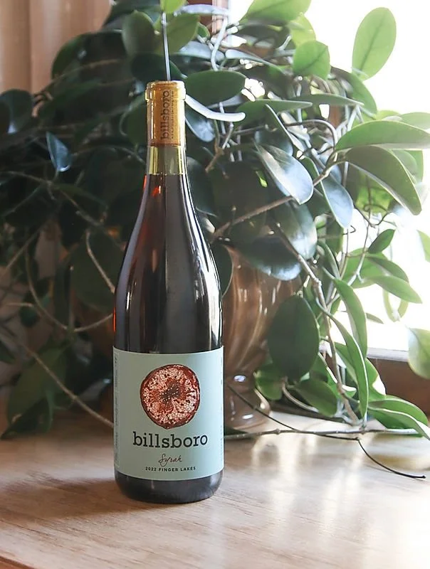 A bottle of Billsboro Spark wine placed on a wooden table in front of a large green leafy plant, with sunlight coming through a window in the background.
