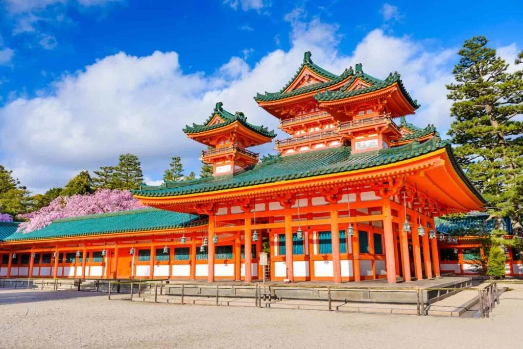 Traditional Japanese shrine with red and white walls, green-tiled roofs, and surrounding trees under a partly cloudy sky.