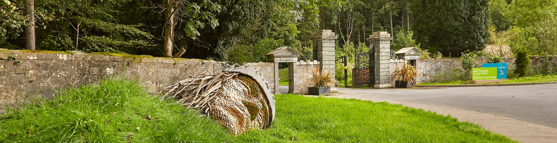 A rolled beehive resting on the grass near a stone wall at the entrance of a park.
