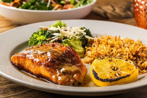 Plate with grilled lemon, cooked salmon with sauce, broccoli with cheese, and a side of noodles.