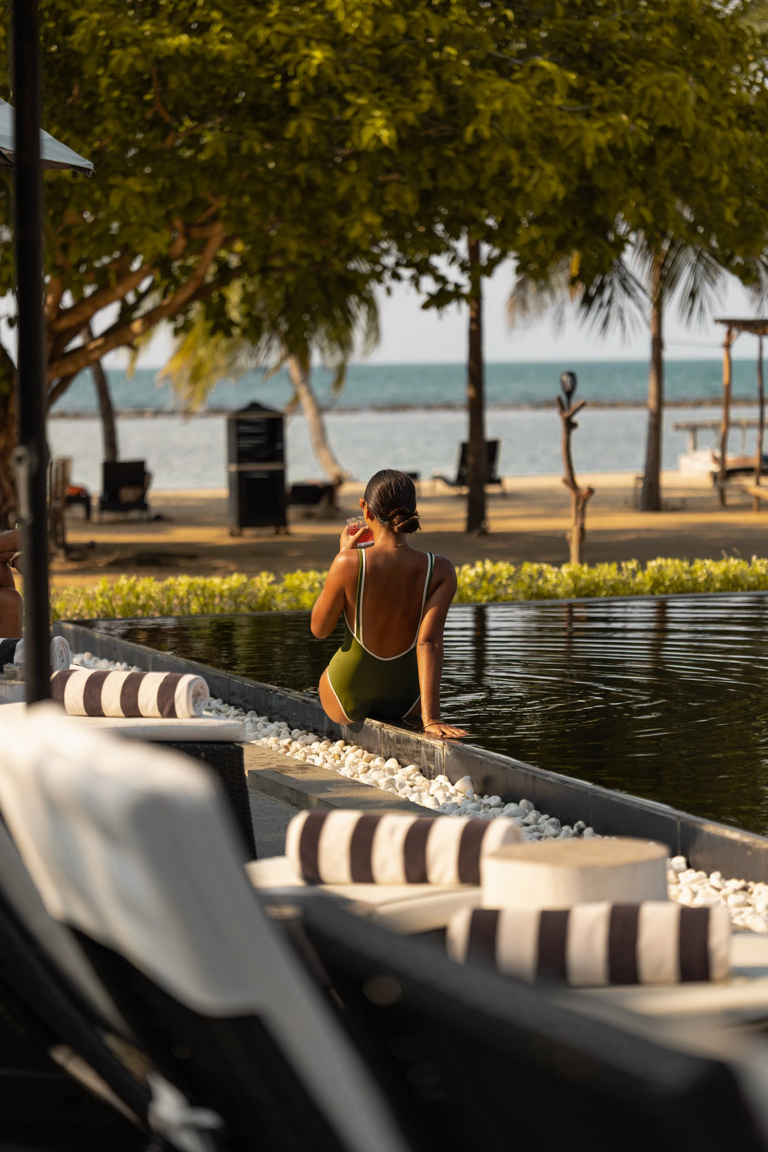 Woman in a one-piece swimsuit sitting by an outdoor pool under trees, with a beach and ocean in the background.