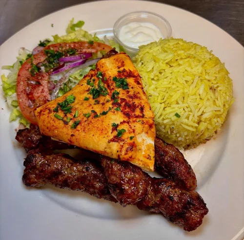 Plate with grilled chicken breast, seasoned rice, grilled beef skewer, fresh salad with tomato and purple onion, and a small container of white sauce.