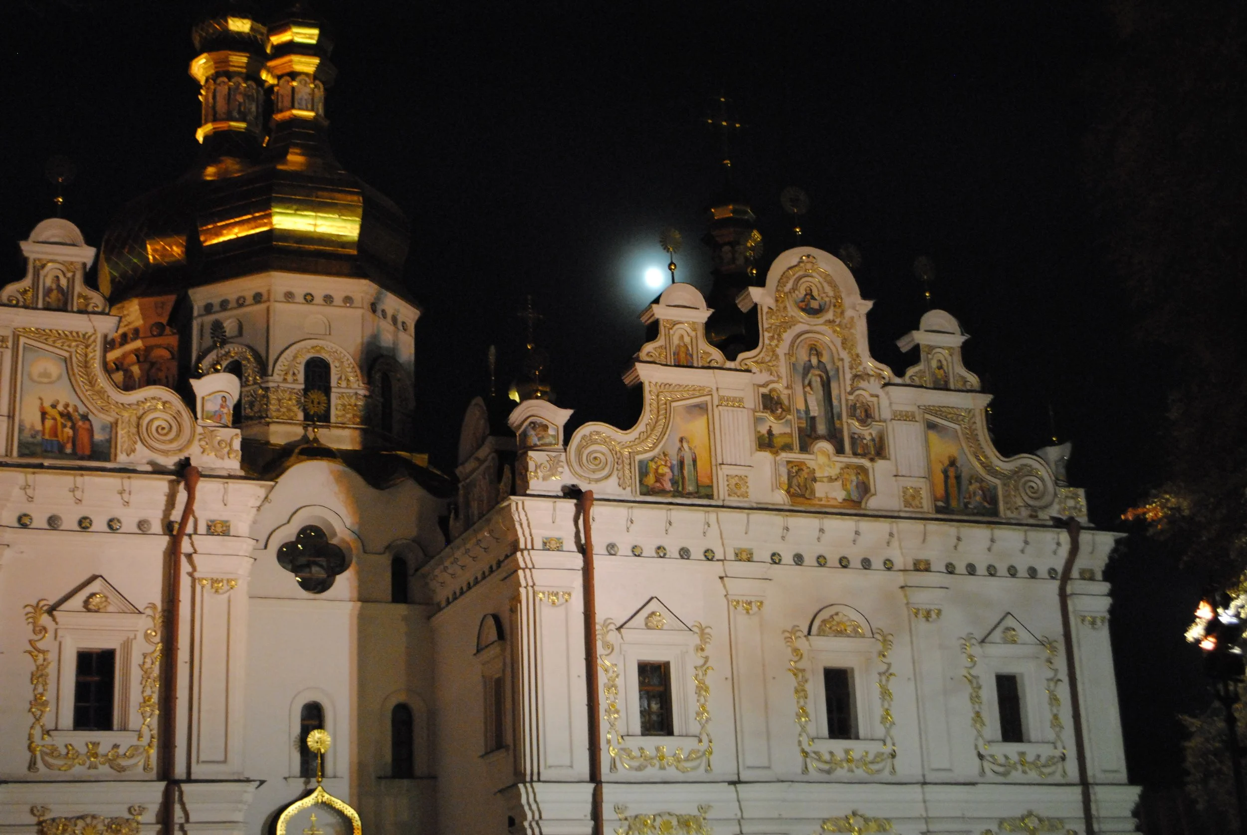 Night view of an ornate, white and gold church with multiple domes and religious icons, illuminated against a dark sky with a bright moon.