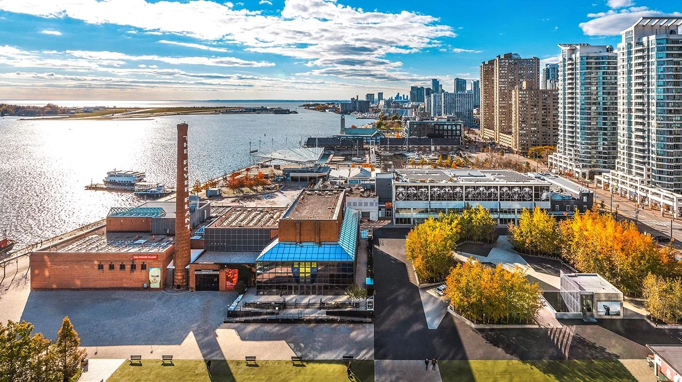 Aerial view of a city waterfront with tall modern buildings, trees with fall foliage, a marina with boats, and a brick power plant with a chimney labeled "The Power Plant" on a sunny day.
