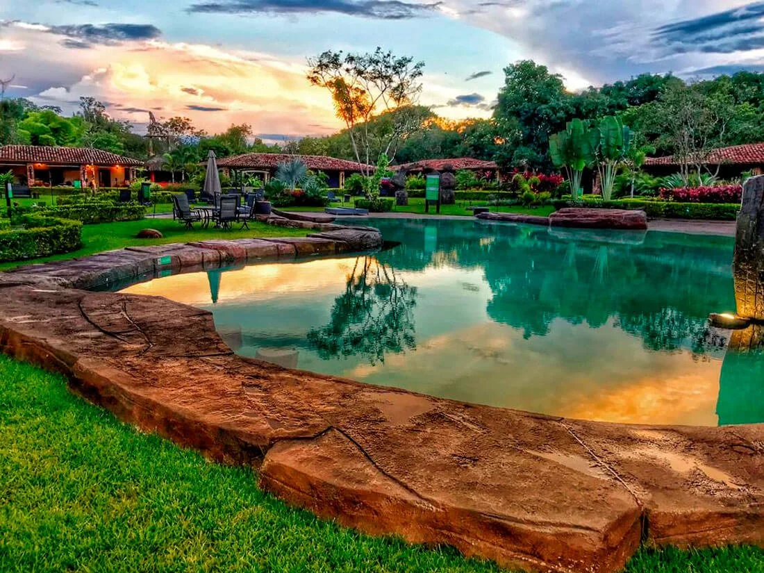 A serene outdoor swimming pool area at sunset, surrounded by lush green trees, colorful flowering plants, and rustic buildings with red-tiled roofs.