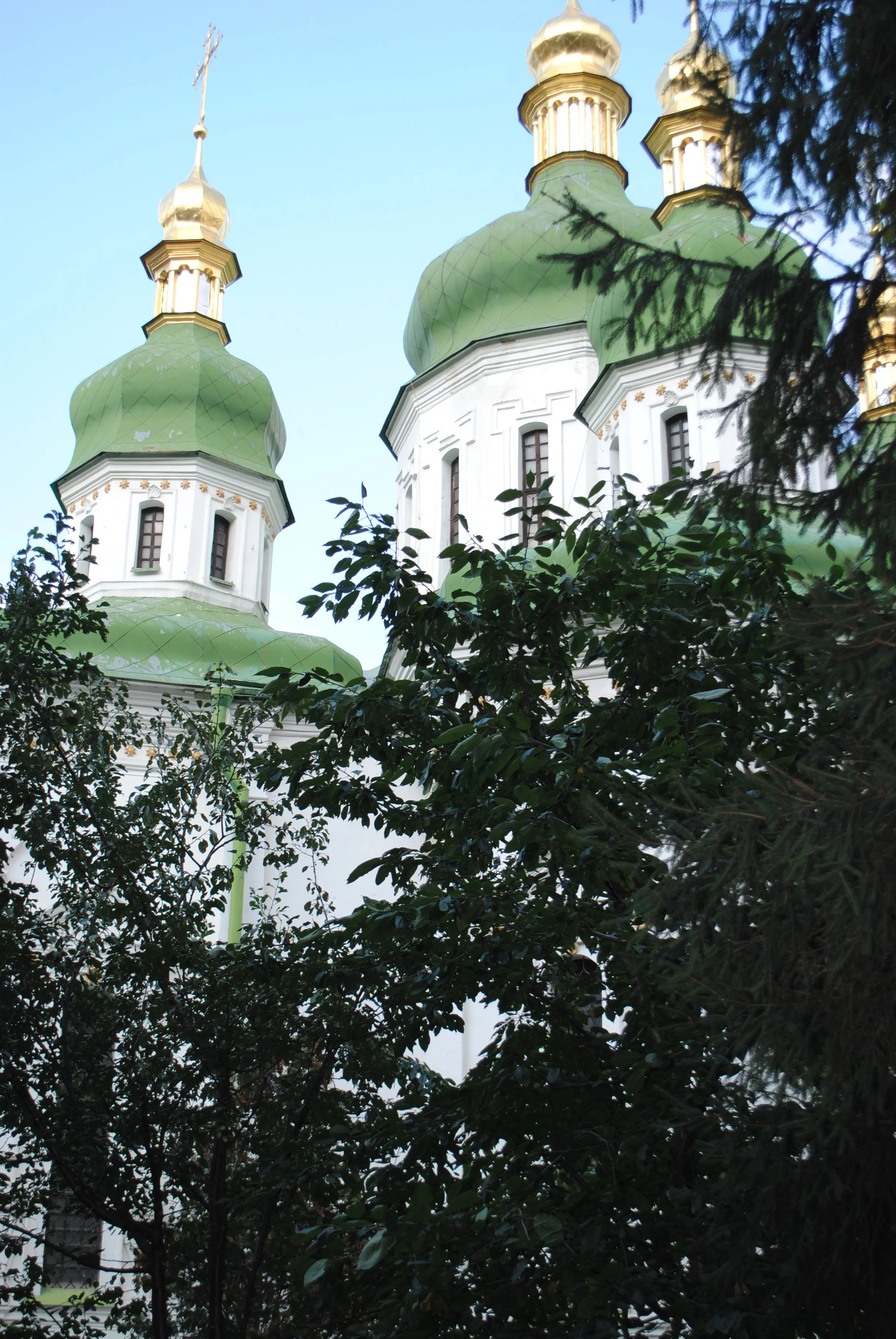 View of a church with green onion-shaped domes and golden crosses, partially obscured by dark green foliage, set against a pale blue sky.