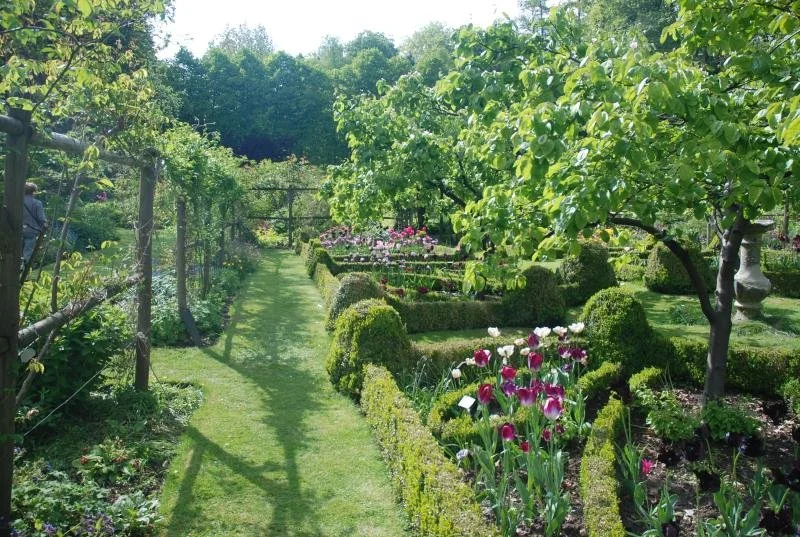 A lush garden with a grass pathway, surrounded by neatly trimmed bushes, colorful flowers, and leafy trees on a sunny day.