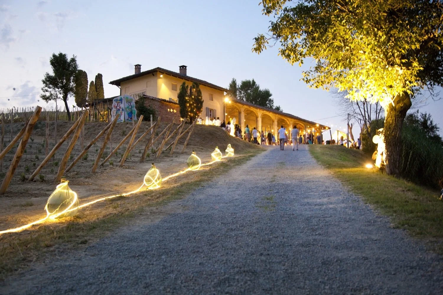 An outdoor evening event with people walking up a gravel path decorated with glowing string lights. The path leads to a large illuminated building with arched openings, surrounded by trees and outdoor decorations.