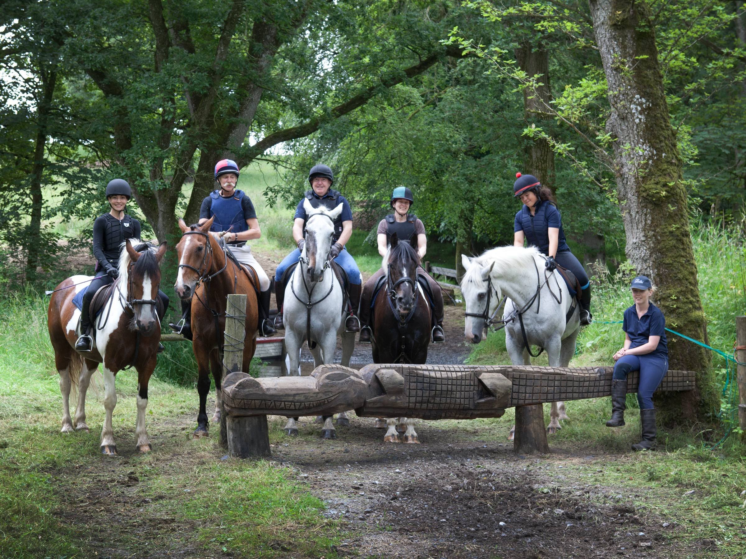A group of people riding horses on a trail through a wooded area, with kids and adults wearing helmets and riding gear, and a woman sitting on a log with a dog nearby.
