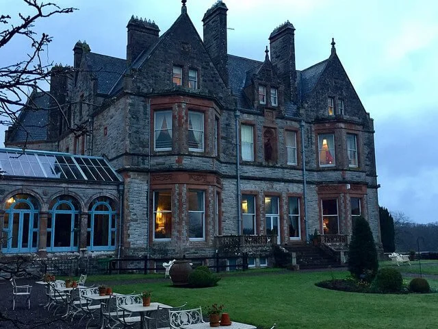 A large, historic stone mansion with multiple towers and windows, surrounded by a well-manicured lawn with outdoor seating and potted plants.