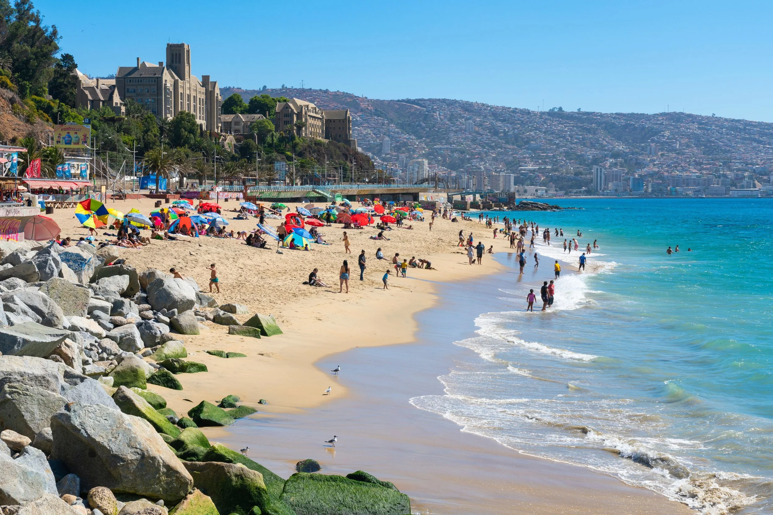 A crowded beach with colorful umbrellas, people swimming and relaxing near the shoreline, rocky foreground, hills with buildings in the background, and a blue sky.