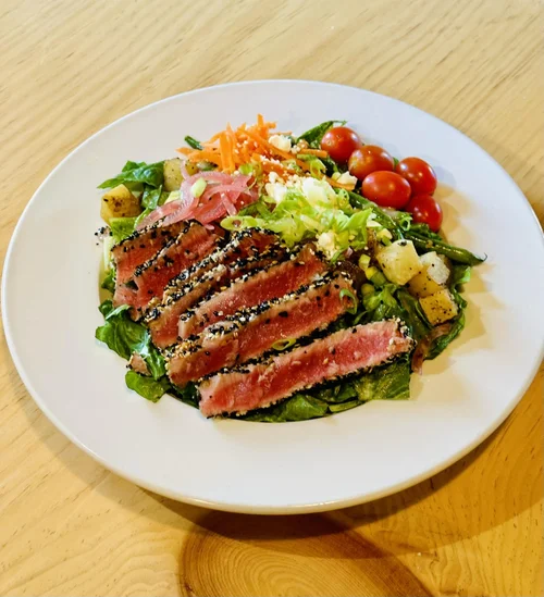 A plate of seared tuna steak on a bed of mixed greens with cherry tomatoes, diced potatoes, shredded carrots, and pickled onions.