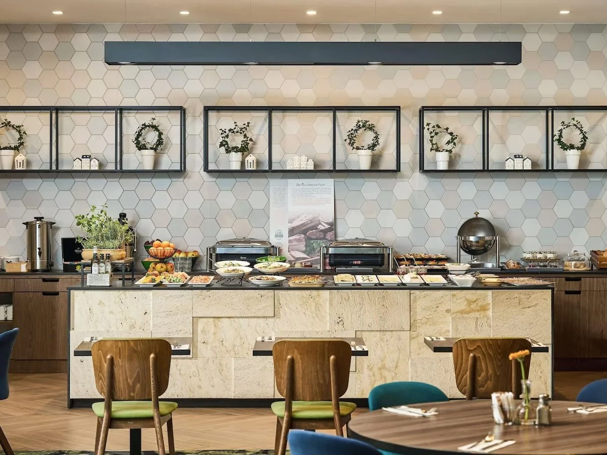 Buffet breakfast spread in a modern dining area with various food items, plates, and utensils, set against a hexagon tiled wall with decorative plants on black metal shelves.