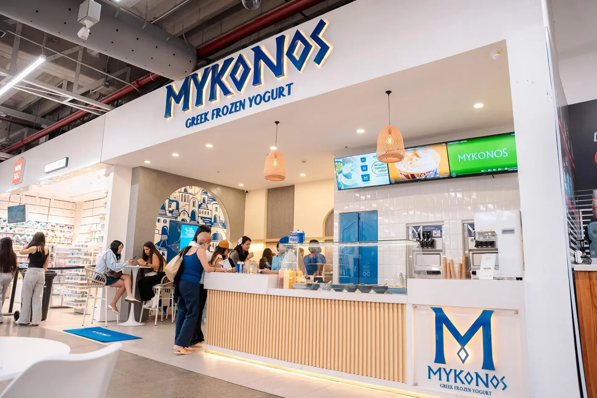 Mykonos Greek frozen yogurt shop inside a shopping mall with customers ordering and eating yogurt. The shop has a modern design with a white counter, digital menu screens, and hanging pendant lights. The signage displays the Mykonos logo and name.