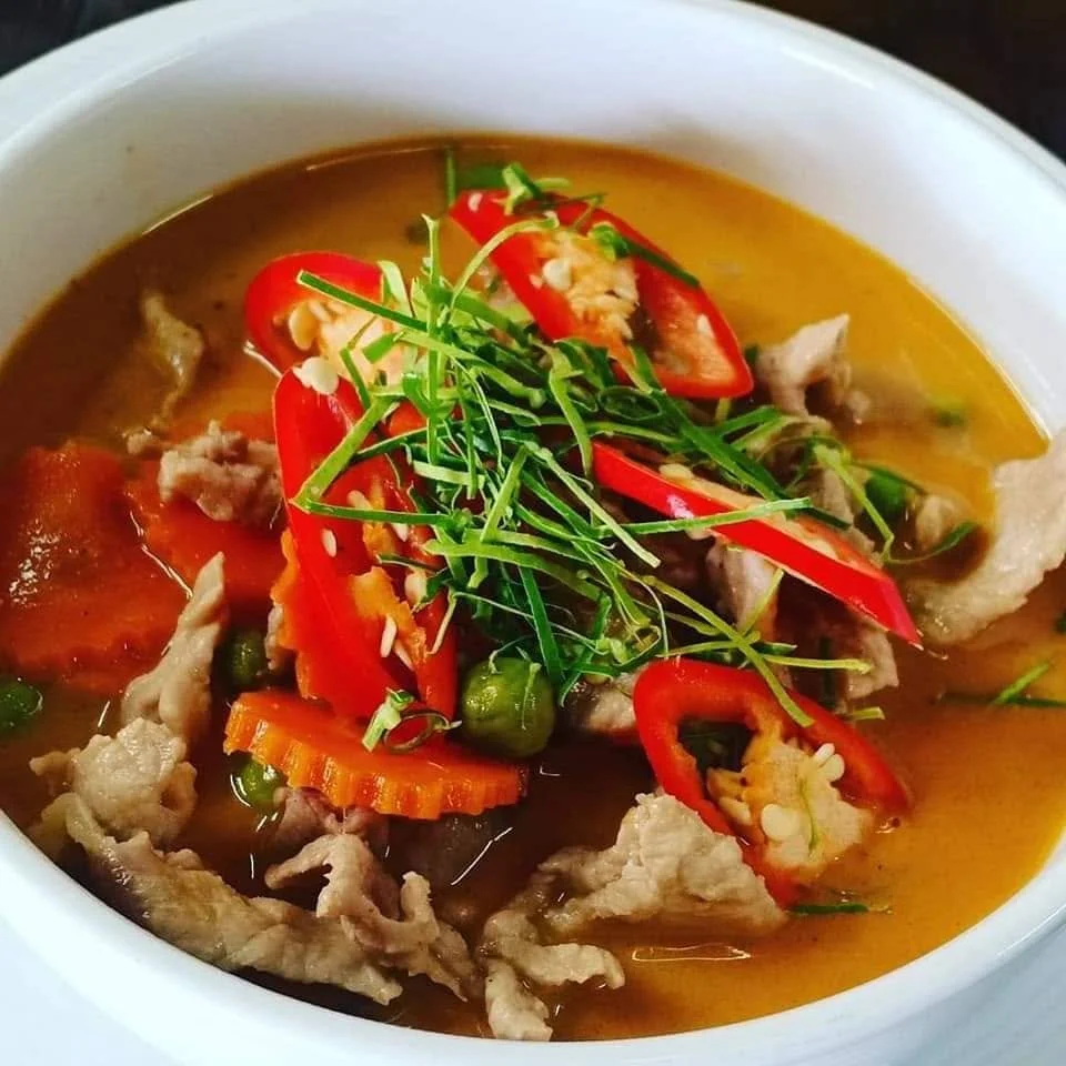 A bowl of soup with sliced red bell peppers, green peas, shredded meat, and garnished with chopped green herbs and sesame seeds.