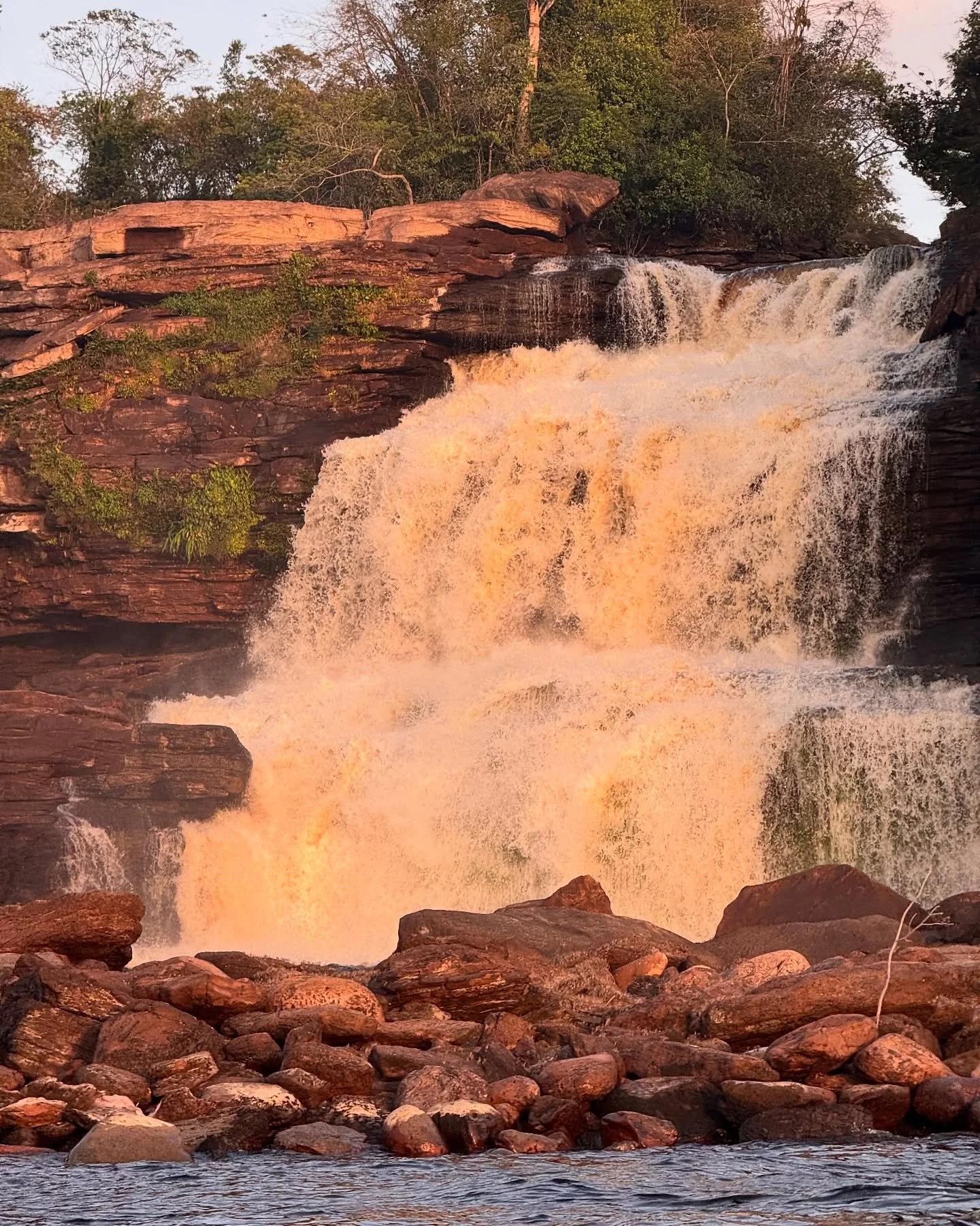 A waterfall cascading over rocks surrounded by green trees during sunset.