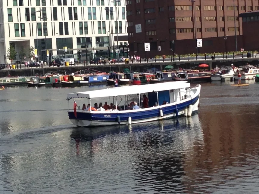 A boat with passengers cruising on a river or canal in an urban area with multiple boats docked along the waterway and tall buildings in the background.