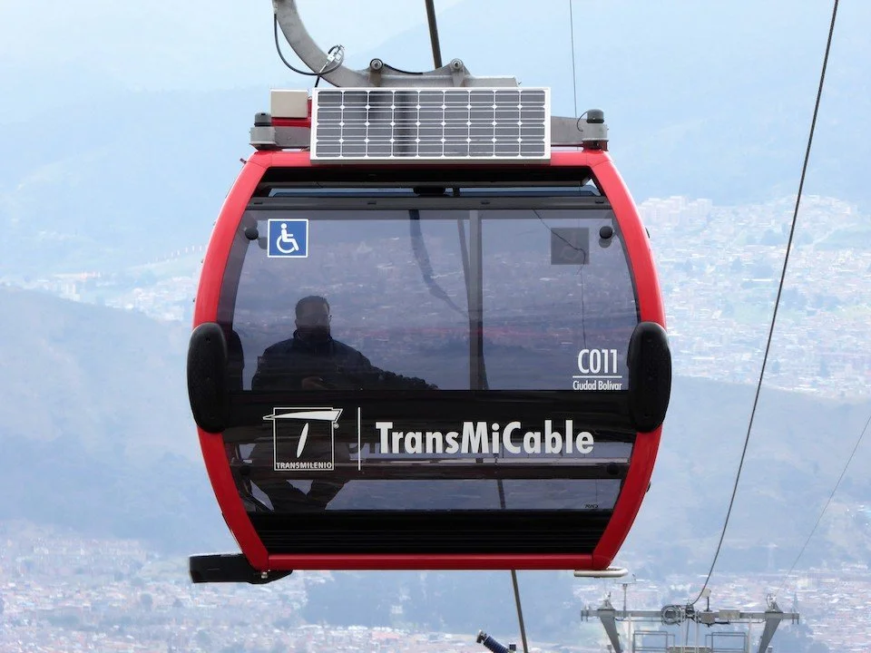 A red cable car with a black window, showing a reflection of a person inside, and a cityscape with mountains in the background. The cable car has a wheelchair accessibility sign and branding for TransMiCable.