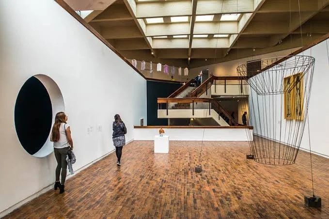 Interior of an art gallery with white walls, wooden floors, and modern artwork, including a wire sculpture and a framed piece. Two women observe the art, and a staircase leads to an upper level with hanging laundry or textiles.