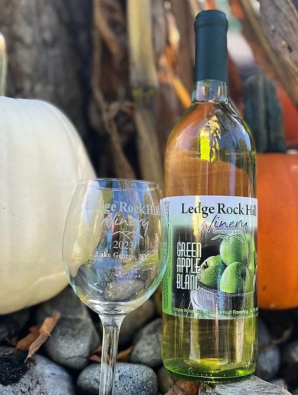 A bottle of green apple white wine from Ledge Rock Hill Winery is positioned next to an empty wine glass outdoors, with decorative pumpkins and rocks in the background.