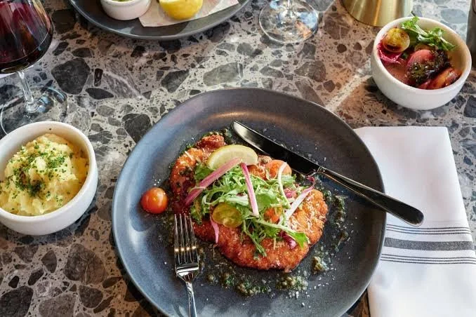 Plated breaded fish fillet garnished with lemon wedge and salad greens, with a fork and knife on a dark plate. Additional dishes include mashed potatoes with herbs, a bowl of cherry tomatoes and greens, a glass of red wine, and a small bowl of salad 