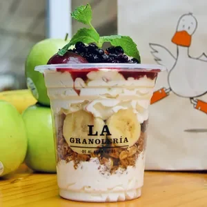A layered apple and berry dessert in a clear cup, topped with mint leaves, with apples and a drawing of a duck on a paper bag in the background.