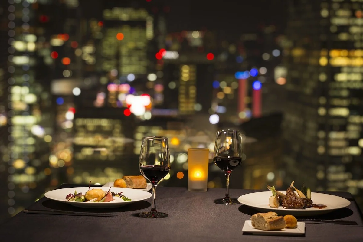A romantic dinner setup on a balcony with two plates of gourmet food, two glasses of red wine, and a candle against a city skyline at night.