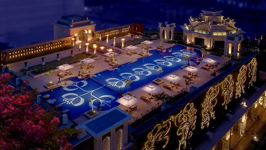 Night view of an ornate rooftop terrace with decorative lighting, a swimming pool with floral patterns illuminated on the water, and luxurious seating areas surrounded by intricate architectural details.